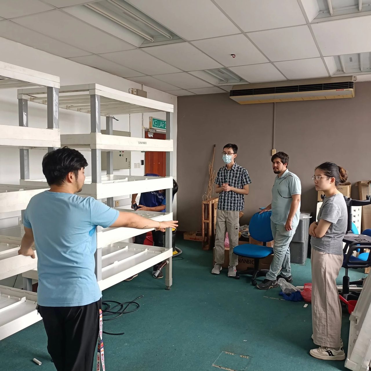 A team member gestures at the empty growing racks while others listen in a room with green carpet