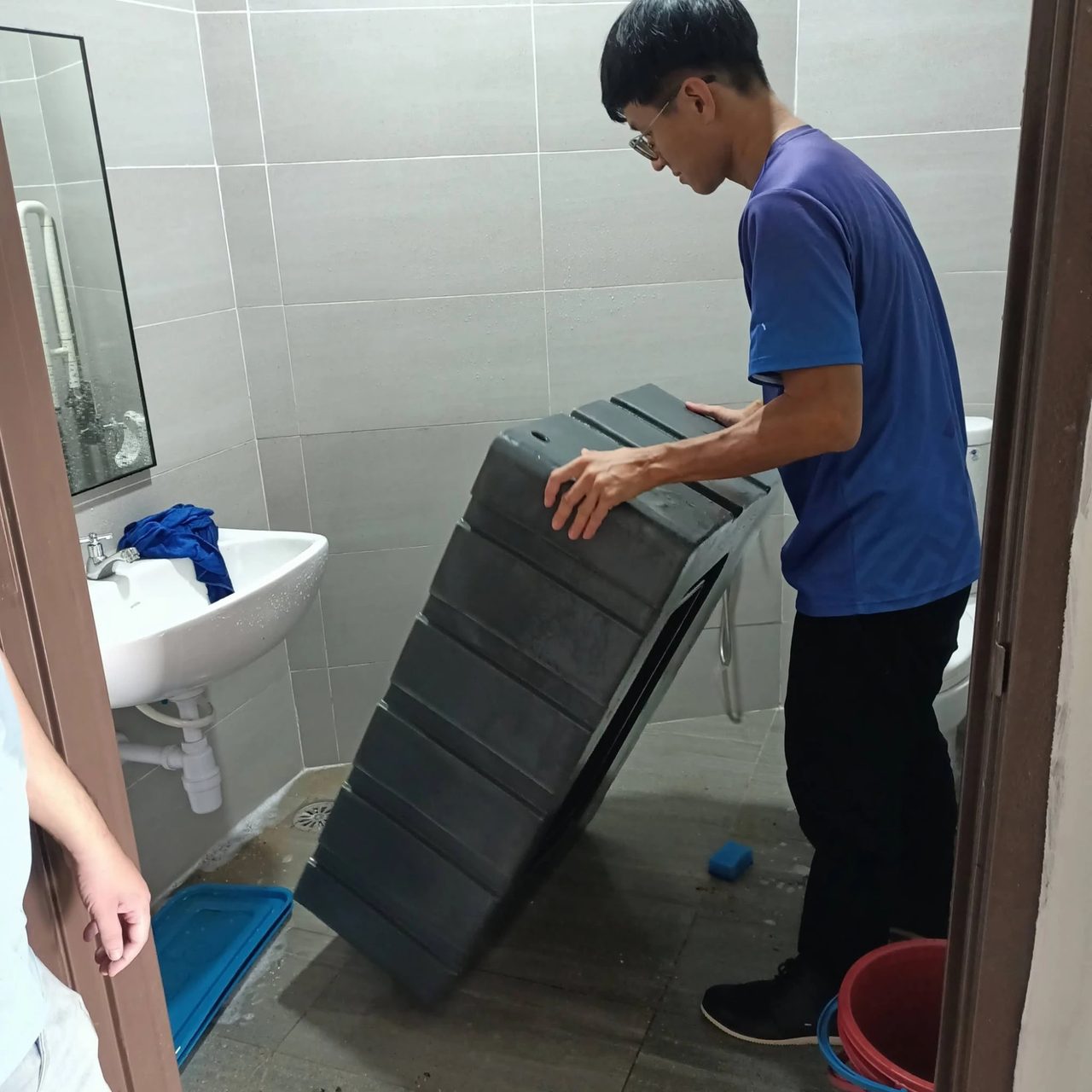 A team member tilts the large grey water tank beside a sink in a washroom to wash it out